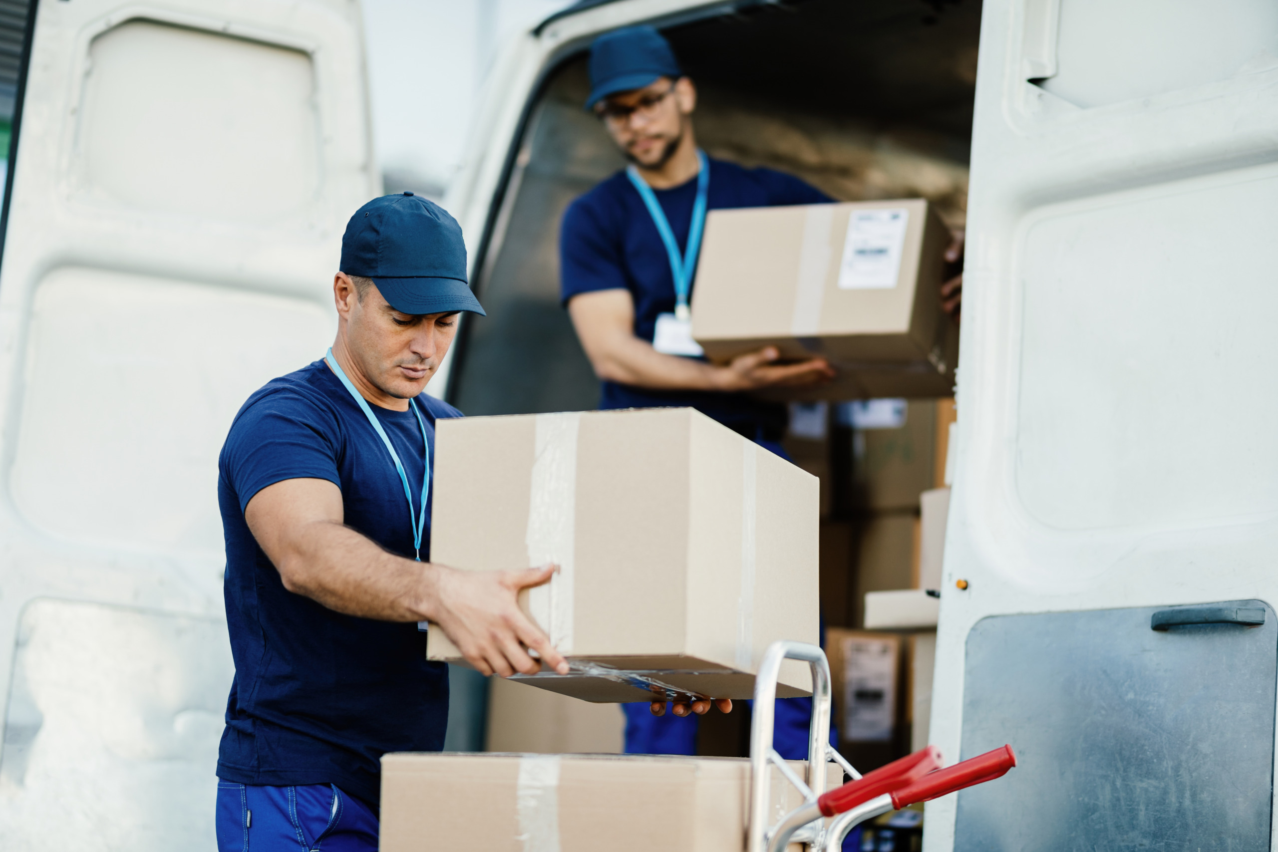 Young delivery man and his coworker unloading boxes from a van. oung-courier-his-colleague-unloading-cardboard-boxes-from-delivery-van-scaled